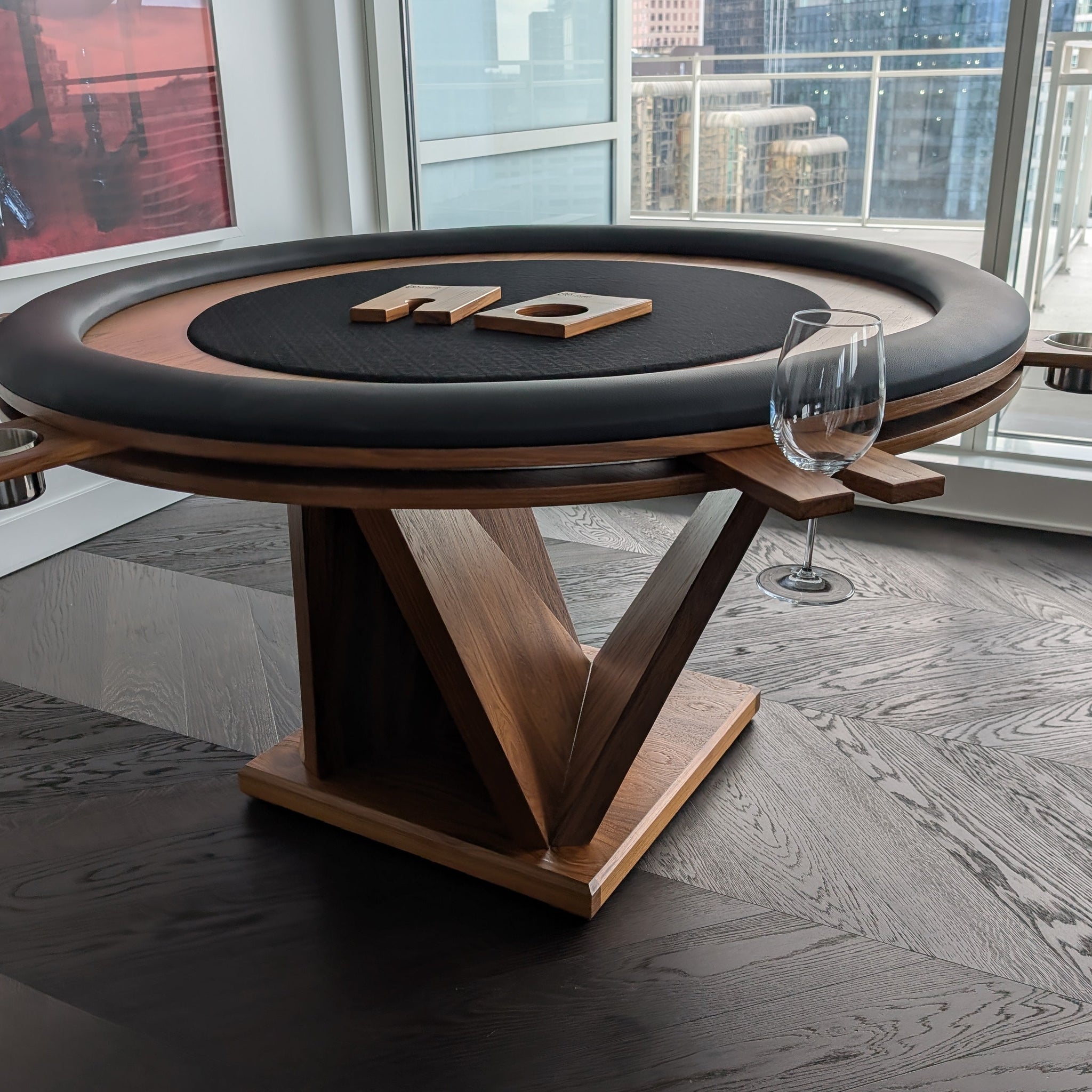 Wooden poker table with black felt top in a room with large windows showing a cityscape.