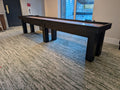 Black shuffleboard table in a room with carpeted floor and large window.