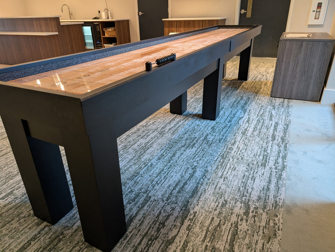 Black shuffleboard table in a room with carpeted floor and large window.