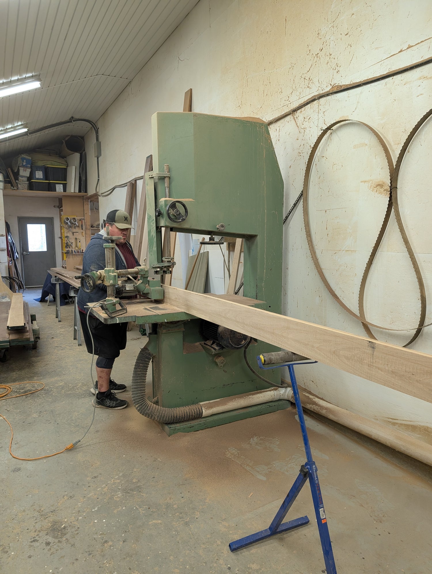 Person operating a band saw in a workshop with wooden planks and tools around.